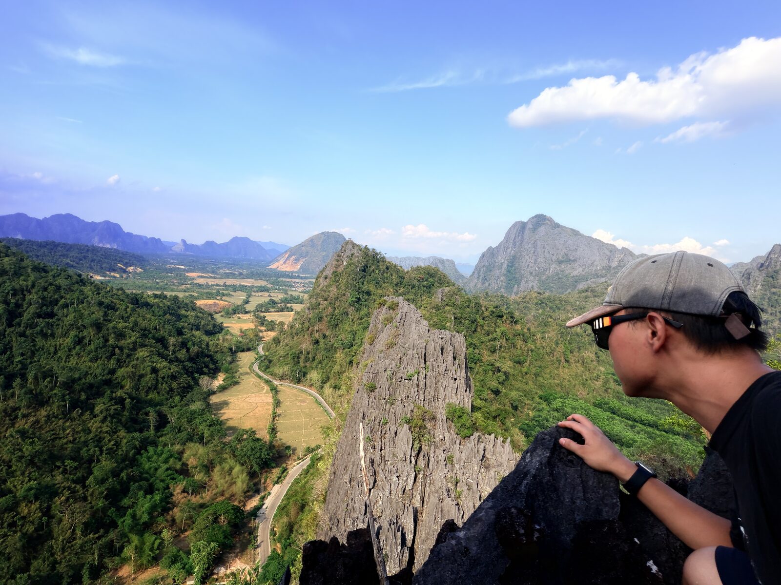 Selfie at a cliff edge on a Vang Vieng summit: the switchback road below, the karst range stretching out in the distance. In that moment you can clearly feel the body’s settled certainty on the edge
